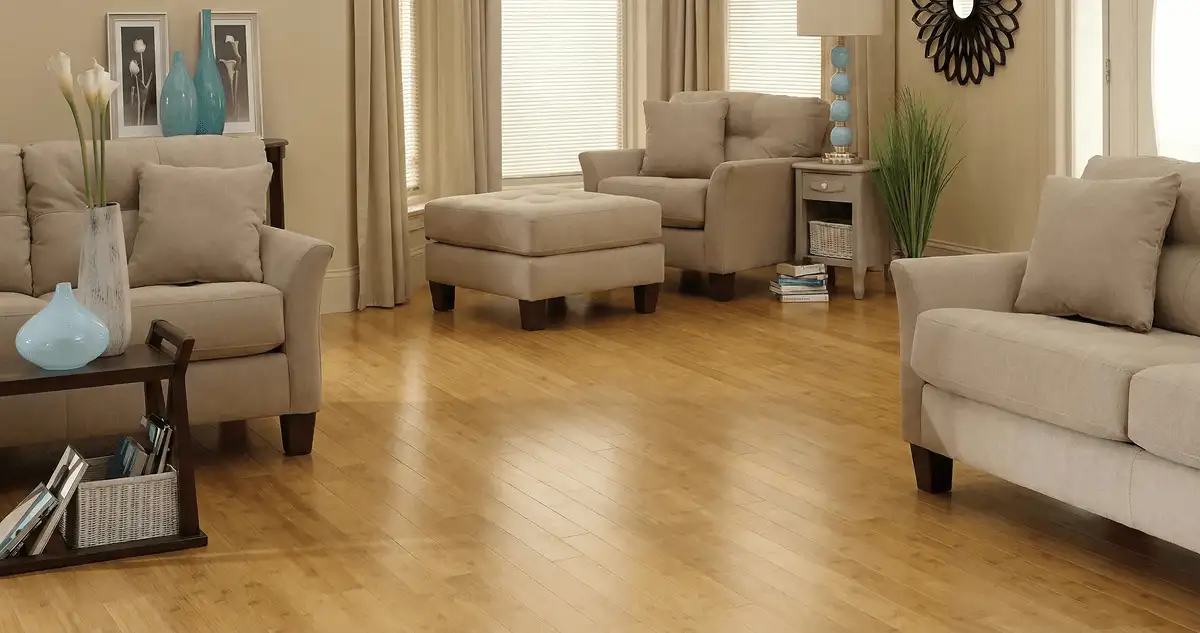 A living room with professionally installed bamboo flooring, beige sofas, a matching ottoman, a side table with a blue lamp, vases, framed pictures, and large windows with blinds and curtains in Allentown, PA